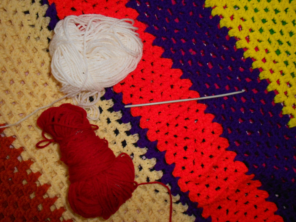 Red and white yarn and a crochet hook resting on colourful crochet fabric.