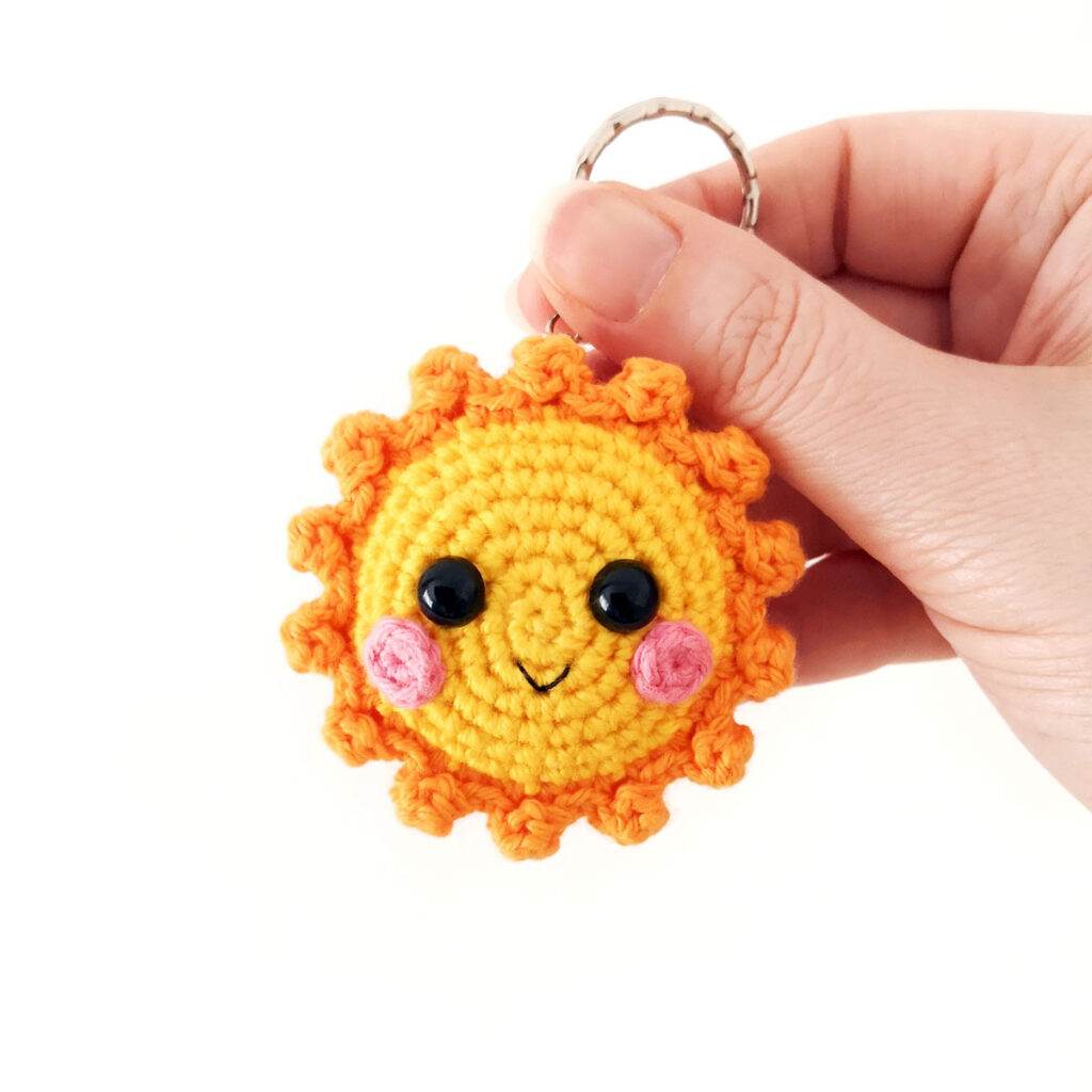 Yellow crochet sun keychain with orange rays and pink cheeks.
