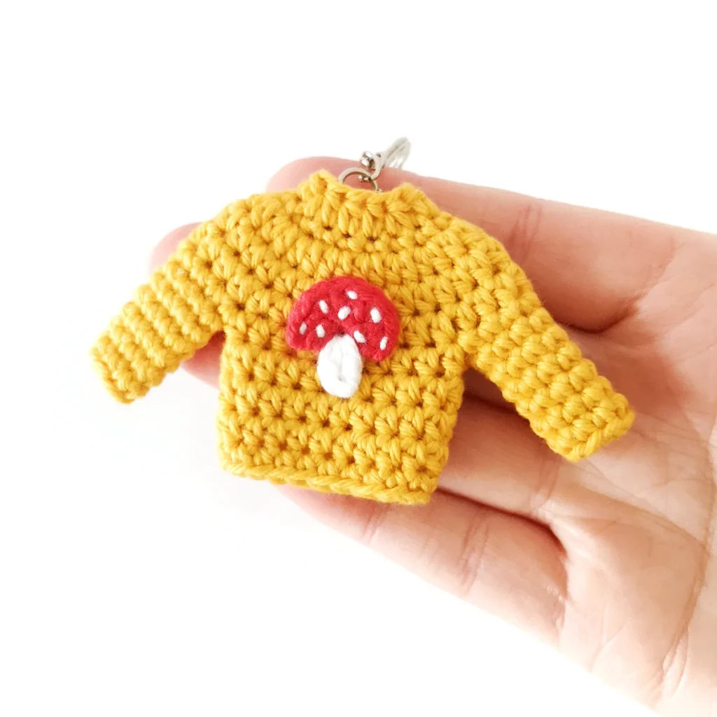 Tiny yellow crochet sweater keychain with a red and white toadstool appliqué.